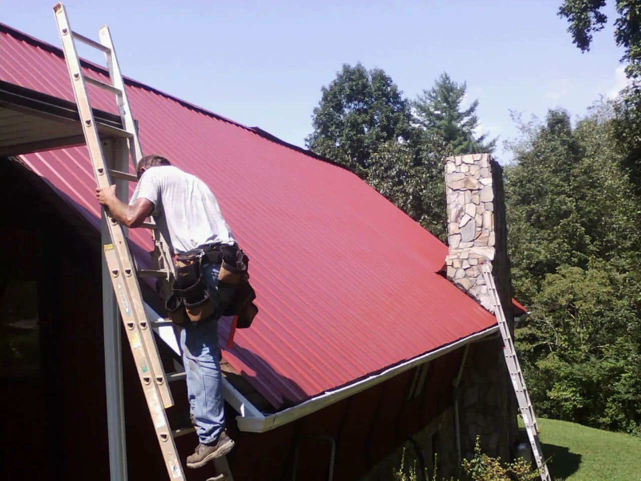 Roofer on a ladder beside a red metal roof and stone chimney — Soffit & Fascia Repair by Roofing Brothers in Elk River